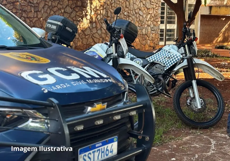 Viatura azul da Guarda Civil Municipal com letras "GCM" no capô e duas motocicletas brancas com padrão quadriculado estacionadas