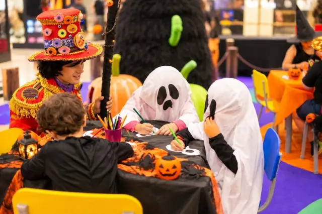 Crianças fantasiadas, duas com lençóis de fantasma, em mesa decorada pintando e desenhando lanternas de Halloween