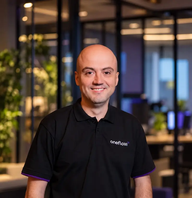 homem calvo sorrindo com camisa polo preta com logotipo 'oneflow', em escritório com divisórias de vidro e plantas ao fundo