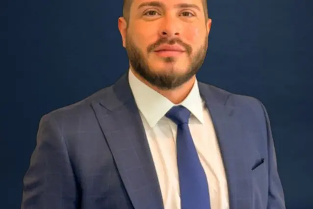 homem adulto de pele clara com barba curta, vestindo terno azul escuro xadrez, camisa branca e gravata azul, em fundo azul liso