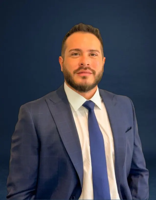 homem adulto de pele clara com barba curta, vestindo terno azul escuro xadrez, camisa branca e gravata azul, em fundo azul liso