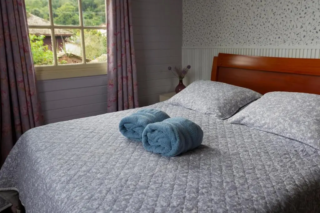 Quarto com cama de casal, colcha cinza e duas toalhas azuis enroladas sobre a cama, janela com vista verde