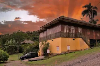 Casarão de madeira elevado sobre base amarela em área verde com céu laranja ao fundo