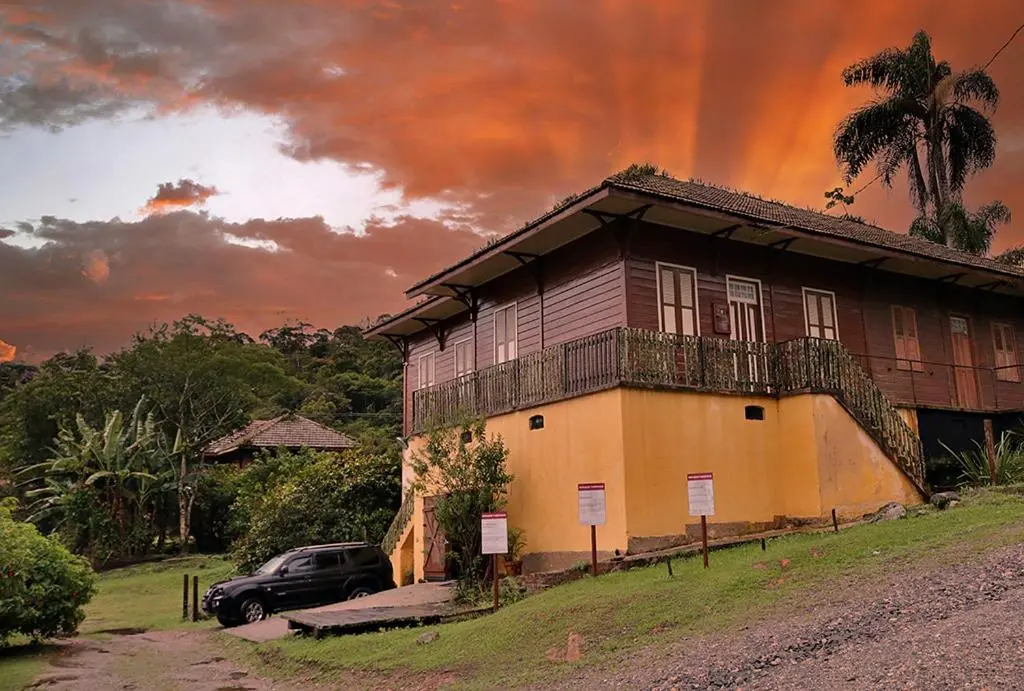 Casarão de madeira elevado sobre base amarela em área verde com céu laranja ao fundo