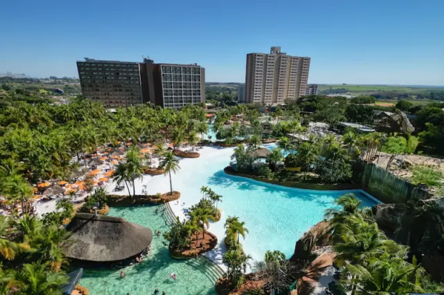 Vista aérea do parque aquático com grandes piscinas, ilhas com palmeiras, espreguiçadeiras e dois prédios de hotel ao fundo