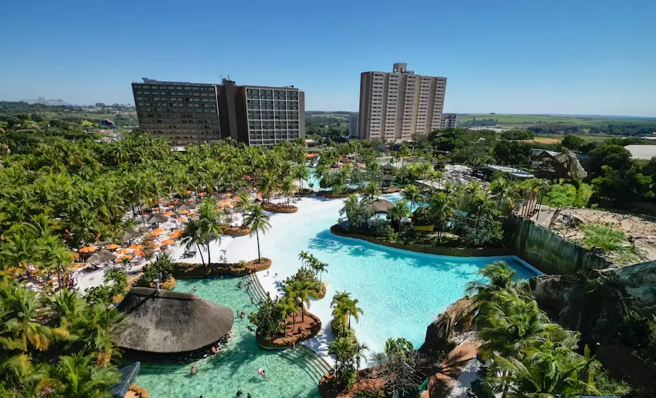 vista aérea de complexo aquático com grandes piscinas, palmeiras, espreguiçadeiras e prédios de hotel ao fundo