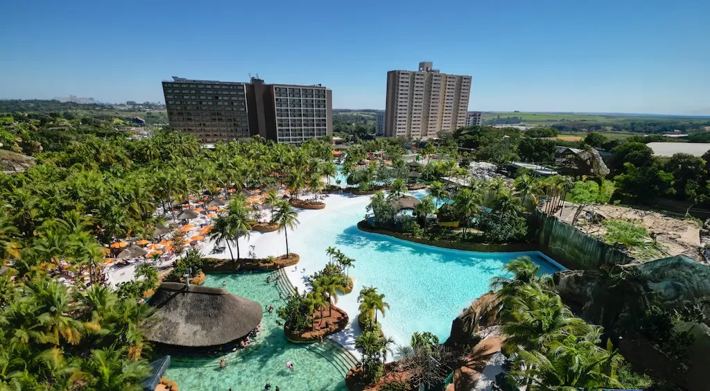 vista aérea de complexo aquático com grandes piscinas, palmeiras, espreguiçadeiras e prédios de hotel ao fundo