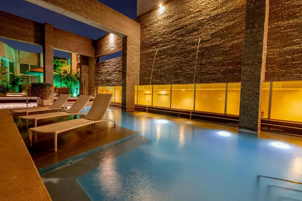Piscina externa com espreguiçadeiras parcialmente submersas junto a muro de pedra iluminado