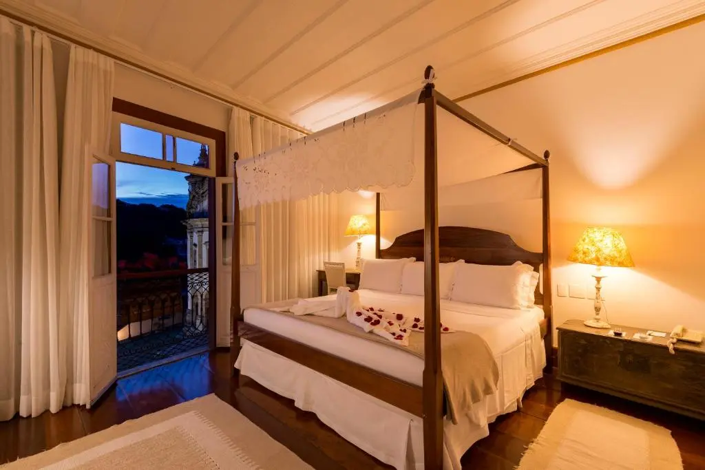 Quarto com cama de dossel, cortinas abertas para varanda e vista de igreja ao entardecer