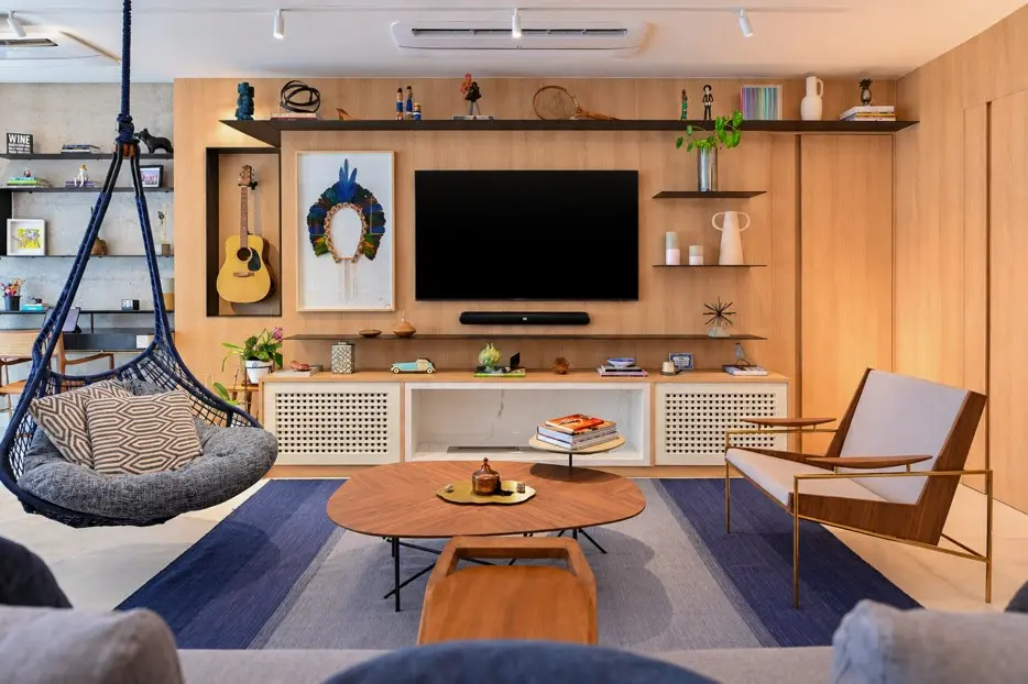 Sala de estar com painel de madeira e televisão, poltrona de madeira, balanço suspenso, mesa de centro redonda e tapete azul