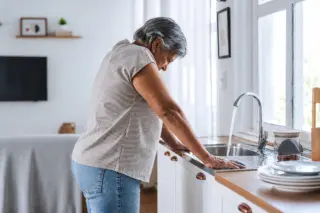 mulher idosa inclinada sobre a pia da cozinha com as mãos apoiadas na bancada