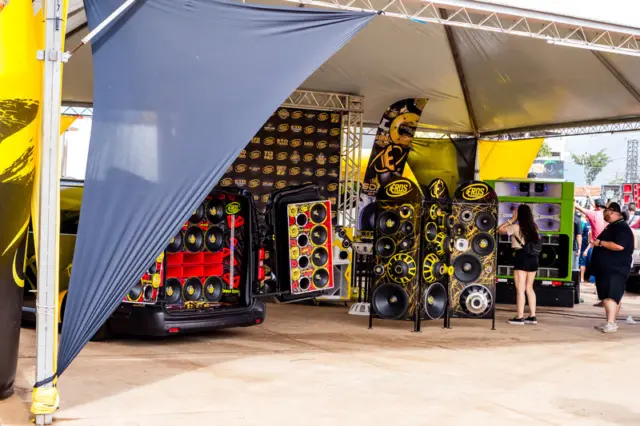 Estande com portas traseiras de veículos abertas exibindo múltiplos alto-falantes e subwoofers, painéis e racks de som sob uma tenda