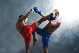 Dois lutadores de Muay Thai trocando chutes altos: um de shorts vermelhos e luvas brancas, outro de shorts azuis e luvas azuis