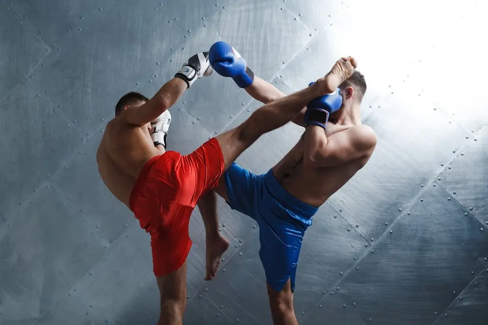 Dois lutadores de Muay Thai trocando chutes altos: um de shorts vermelhos e luvas brancas, outro de shorts azuis e luvas azuis