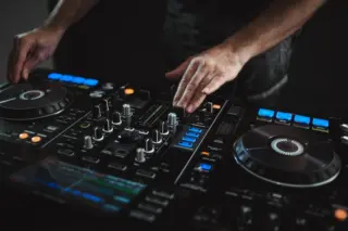 Mãos de um DJ ajustando controles em uma mesa de mixagem com botões iluminados
