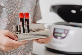 Duas amostras de sangue em tubos com tampa vermelha em suporte metálico, com centrífuga desfocada ao fundo