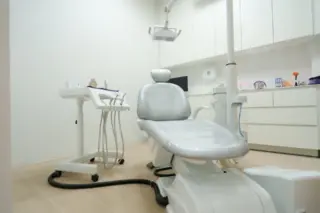 Sala de atendimento odontológico com cadeira reclinável cinza no centro, suportes de instrumentos e armários brancos ao fundo
