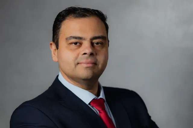 Homem adulto em retrato profissional, vestindo terno escuro, camisa azul clara e gravata vermelha, posando com braços cruzados contra fundo cinza