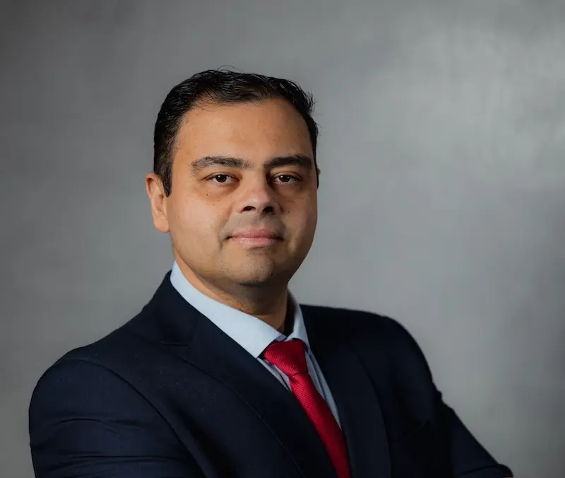Homem adulto em retrato profissional, vestindo terno escuro, camisa azul clara e gravata vermelha, posando com braços cruzados contra fundo cinza