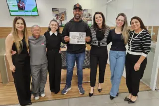 Sete pessoas em pé dentro de uma agência, homem ao centro segurando um papel impresso e equipe posando ao lado