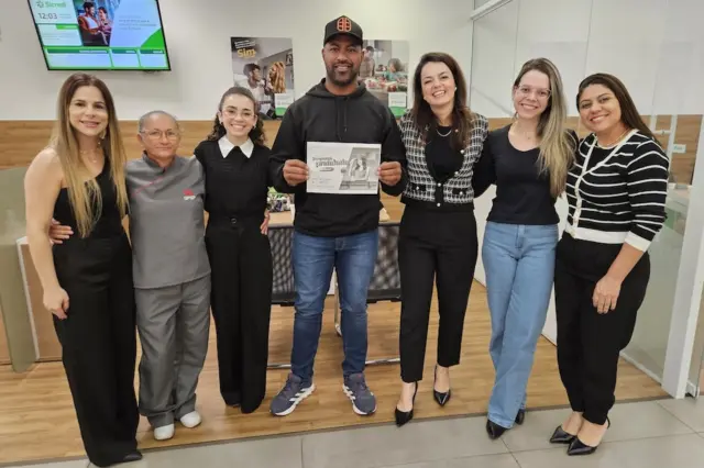 Sete pessoas em pé dentro de uma agência, homem ao centro segurando um papel impresso e equipe posando ao lado