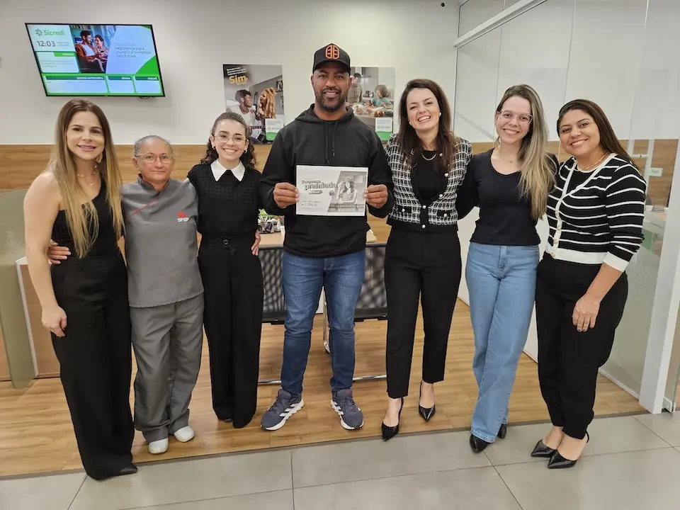Sete pessoas em pé dentro de uma agência, homem ao centro segurando um papel impresso e equipe posando ao lado