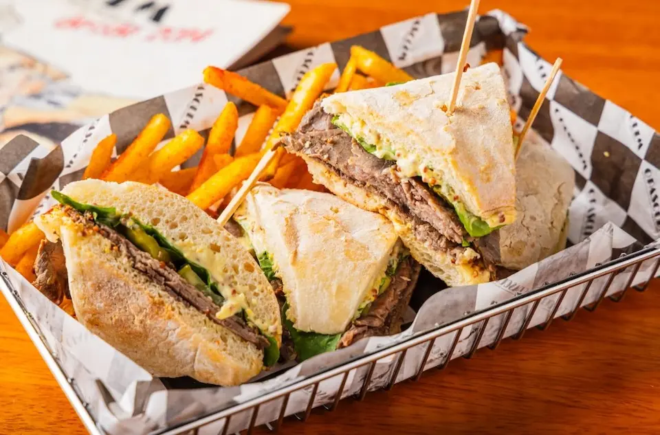 pães ciabatta cortados em triângulos recheados com fatias de carne, rúcula e maionese, servidos em cesta com batatas fritas