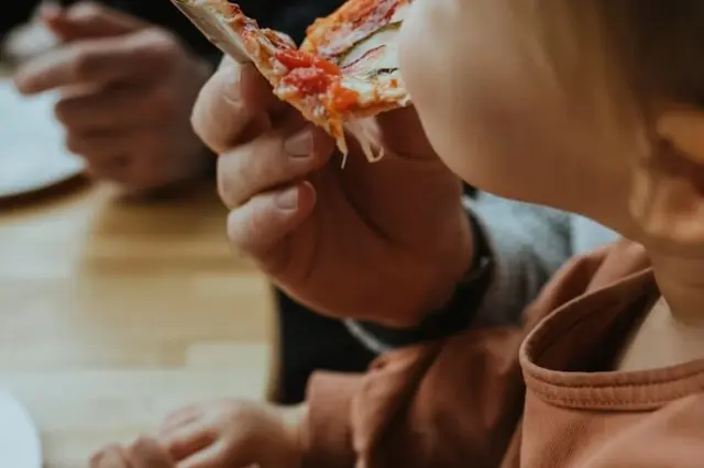 Criança de perfil comendo uma fatia de pizza segurada por uma mão adulta sobre mesa