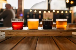 quatro copos pequenos com diferentes cervejas alinhados sobre uma tábua de madeira em uma mesa de bar
