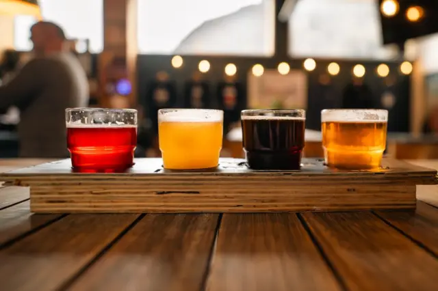 quatro copos pequenos com diferentes cervejas alinhados sobre uma tábua de madeira em uma mesa de bar