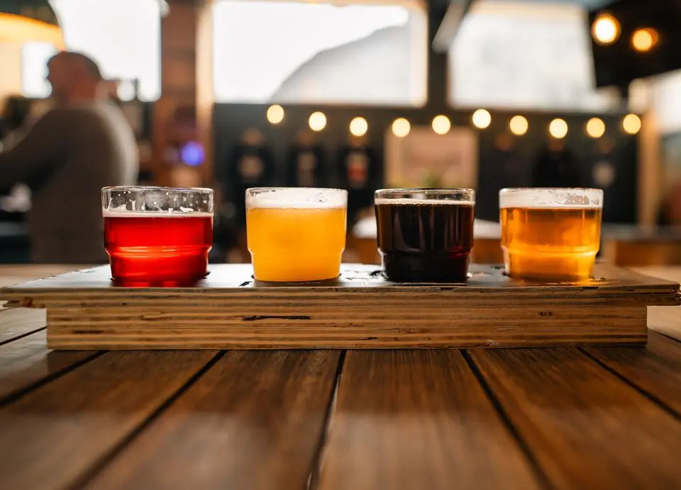 quatro copos pequenos com diferentes cervejas alinhados sobre uma tábua de madeira em uma mesa de bar