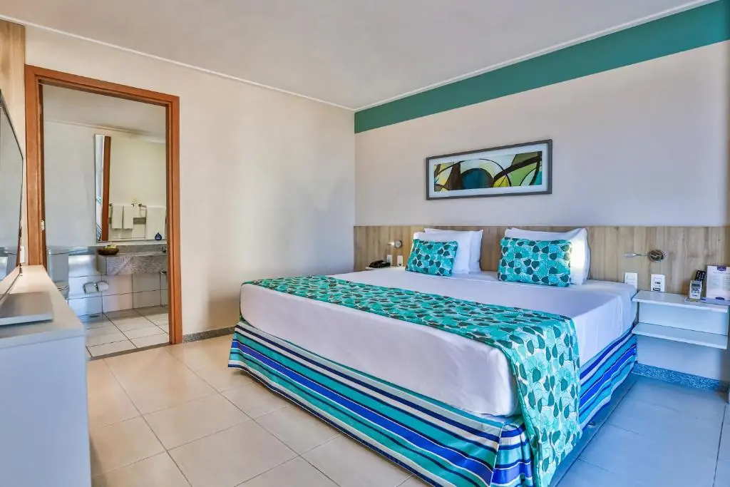 quarto com cama de casal e decoração em tons de azul e verde, banheiro visível pela porta