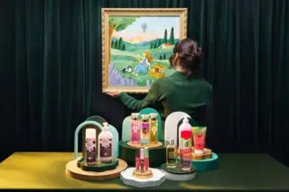 Pessoa de costas segurando um quadro com pintura de paisagem; frascos e embalagens coloridos de cosméticos dispostos sobre pedestais em uma mesa, com cortina verde ao fundo
