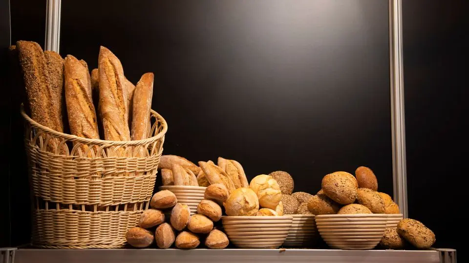 Cesta de vime com baguetes, tigelas com pães variados e pequenos pães sobre balcão, fundo preto