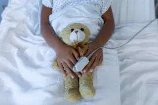 criança deitada em cama de hospital com ursinho de pelúcia no colo e sensor de oximetria preso a um dedo