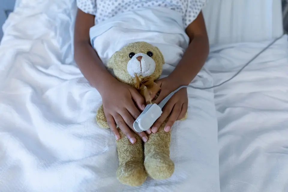 criança deitada em cama de hospital com ursinho de pelúcia no colo e sensor de oximetria preso a um dedo