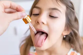 Menina de cabelos castanhos com a língua para fora enquanto um conta-gotas com tampa dourada pinga líquido sobre a língua