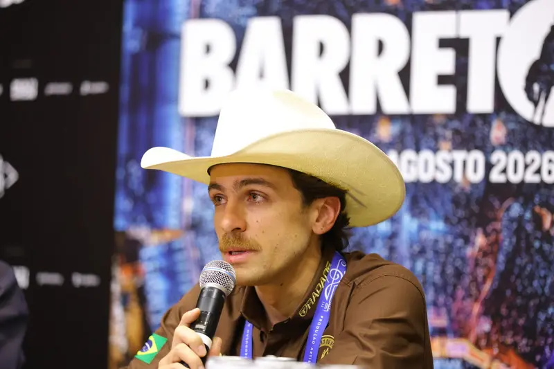 homem usando chapéu branco de cowboy segurando microfone em painel com fundo que exibe a palavra 'BARRETOS'