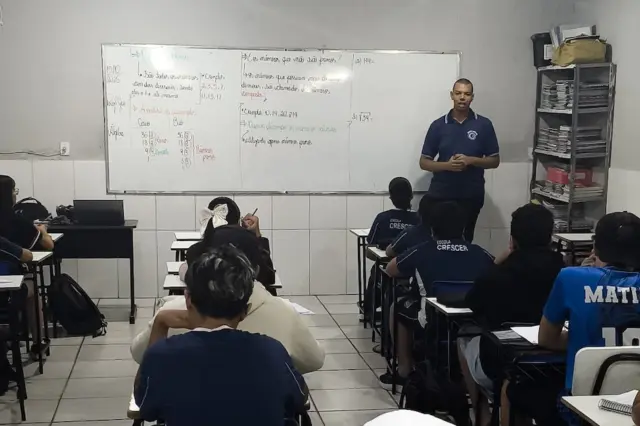 Professor de pé à frente de quadro branco com alunos sentados de costas em carteiras