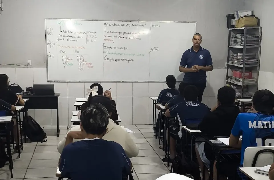 Professor de pé à frente de quadro branco com alunos sentados de costas em carteiras