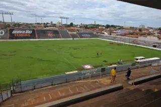 Vista interna do estádio com gramado irregular, degraus de arquibancada em primeiro plano, algumas pessoas no campo e um micro-ônibus junto à lateral; casas e céu nublado ao fundo