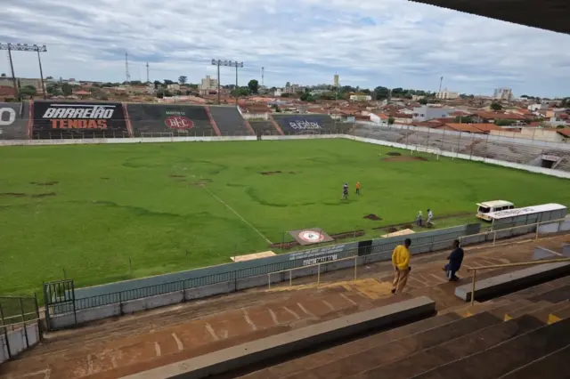 Vista interna do estádio com gramado irregular, degraus de arquibancada em primeiro plano, algumas pessoas no campo e um micro-ônibus junto à lateral; casas e céu nublado ao fundo