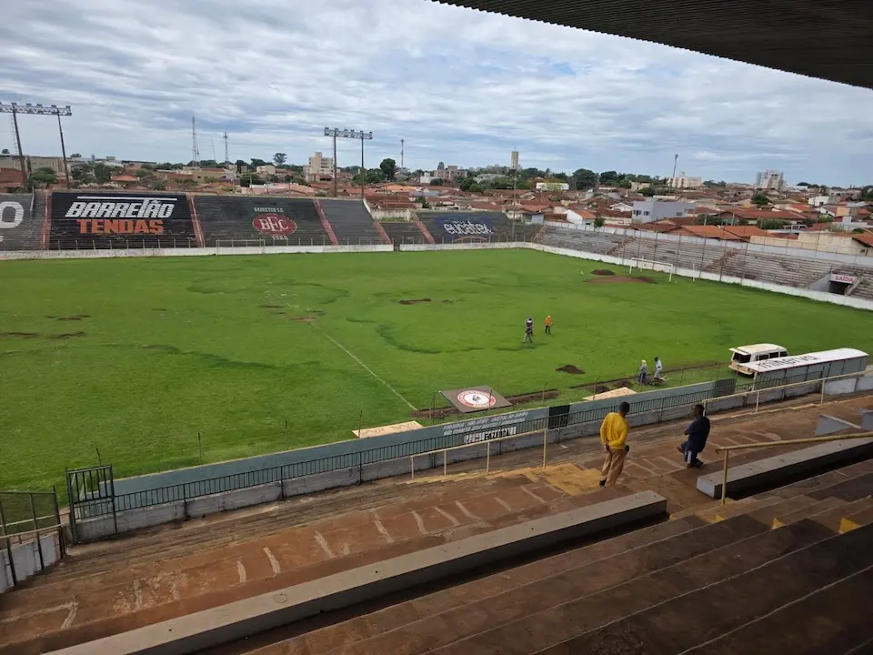Vista interna do estádio com gramado irregular, degraus de arquibancada em primeiro plano, algumas pessoas no campo e um micro-ônibus junto à lateral; casas e céu nublado ao fundo