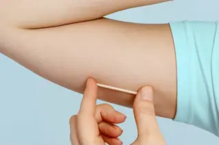 Mão segurando um aplicador junto à face interna do braço de uma pessoa com camiseta azul-clara