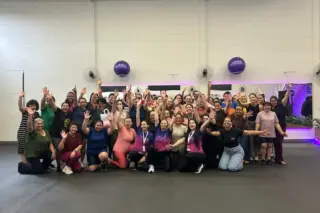 Grupo de mulheres reunidas em academia, a maioria sorrindo e acenando, com bolas de ginástica fixadas na parede e iluminação roxa ao fundo