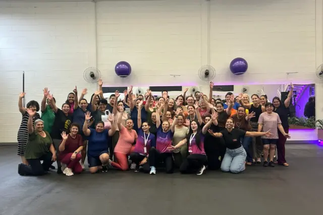 Grupo de mulheres reunidas em academia, a maioria sorrindo e acenando, com bolas de ginástica fixadas na parede e iluminação roxa ao fundo