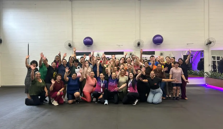 Grupo de mulheres reunidas em academia, a maioria sorrindo e acenando, com bolas de ginástica fixadas na parede e iluminação roxa ao fundo