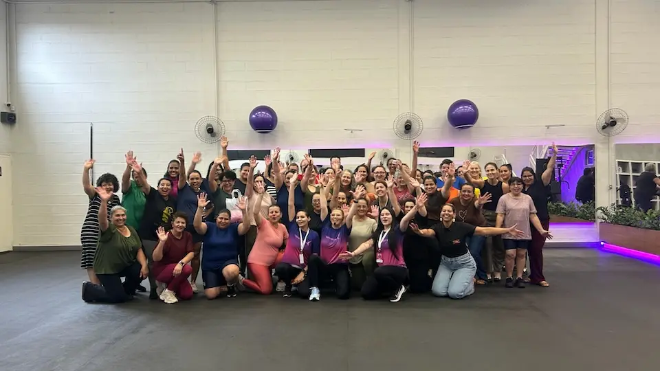 Grupo de mulheres reunidas em academia, a maioria sorrindo e acenando, com bolas de ginástica fixadas na parede e iluminação roxa ao fundo