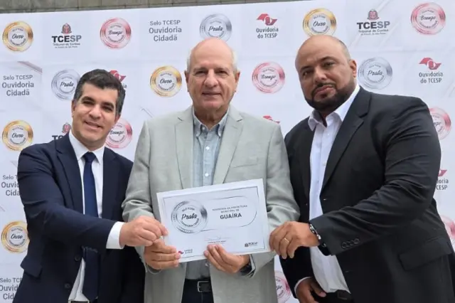 Três homens de terno seguram um certificado diante de um painel com logos e selos do TCE-SP