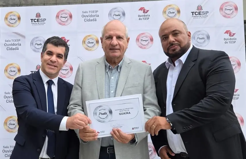 Três homens de terno seguram um certificado diante de um painel com logos e selos do TCE-SP